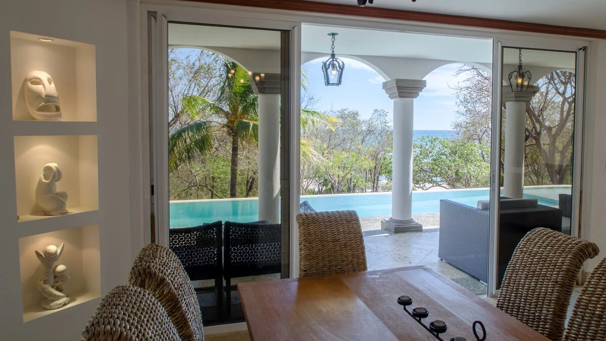 A dining area with woven chairs and a wooden table opens through glass doors toward a pool, columns, trees, and blue water beyond.