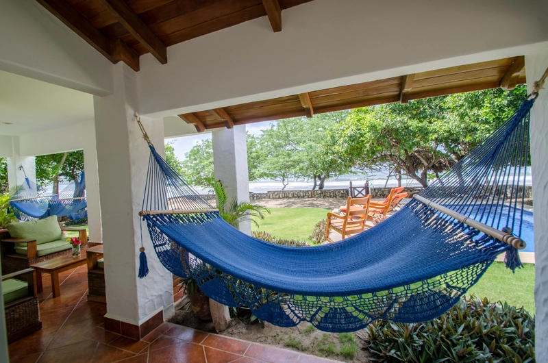 A cozy outdoor patio features a vibrant blue hammock strung between white columns. There are comfortable wooden chairs and a garden view with lush green trees and a well-kept lawn 