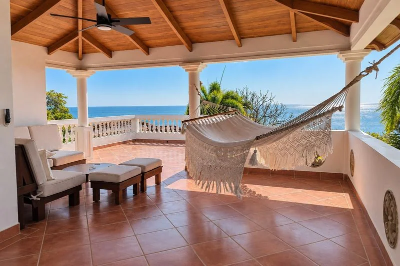 A covered terracotta-tiled terrace with a hanging hammock, lounge seating, wooden ceiling, and ocean visible beyond the balustrade.