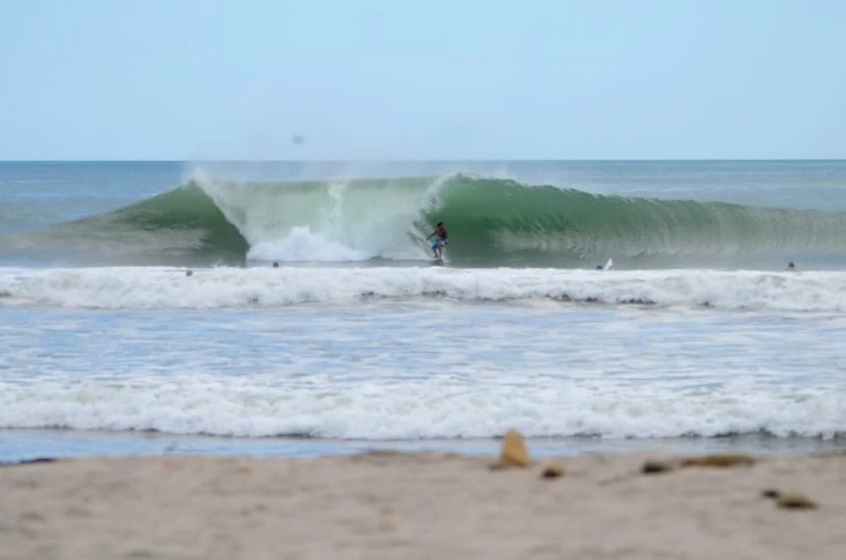A surfer rides a breaking wave offshore beyond a sandy beach.