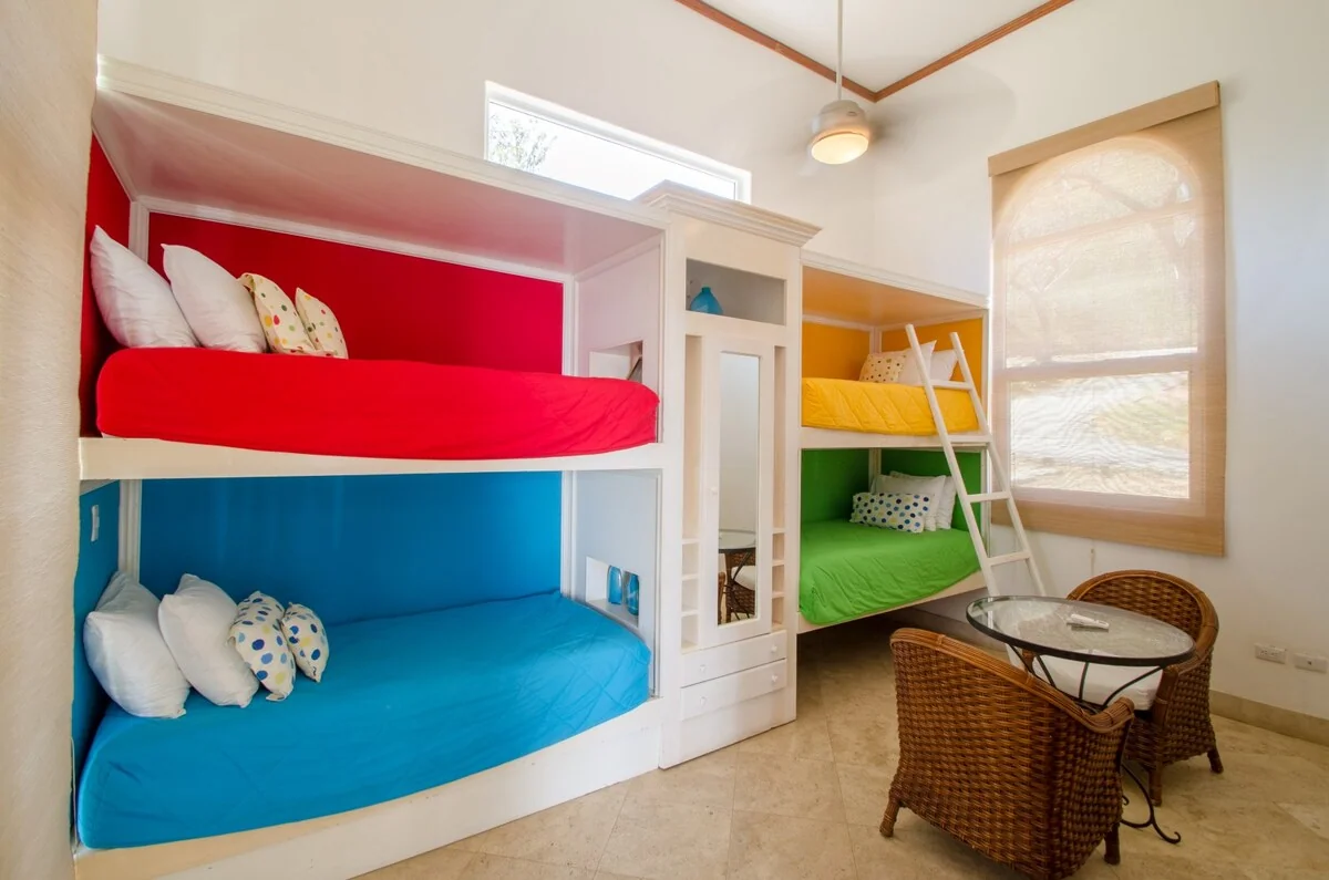 A bright room contains colorful bunk beds, wicker chairs around a glass table, and a large window.