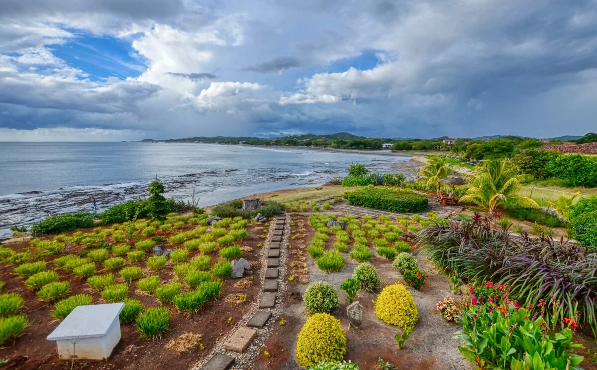 A landscaped coastal garden with flowering plants and a central stone path overlooks a calm shoreline beneath dramatic clouds.