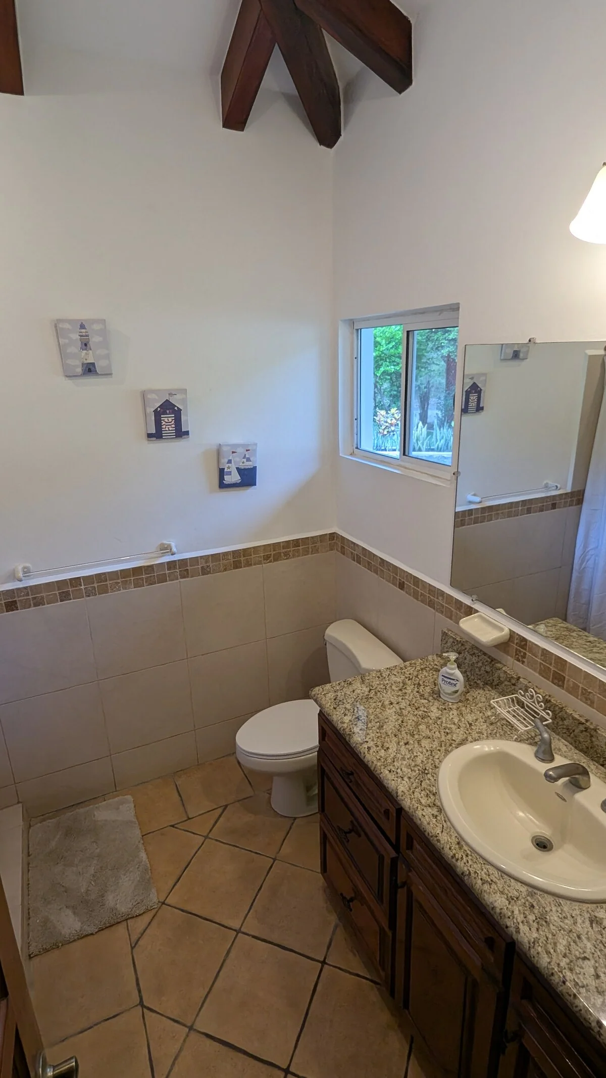 A tiled bathroom with a granite vanity, sink, toilet, wall mirror, small window, and exposed wooden ceiling beams.