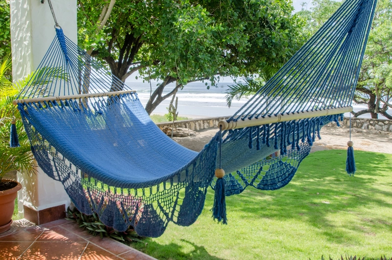 The image shows a blue hammock hanging in a garden. The garden is lush and green, with several trees providing shade. In the background, there is a view of a beach and the ocean. T