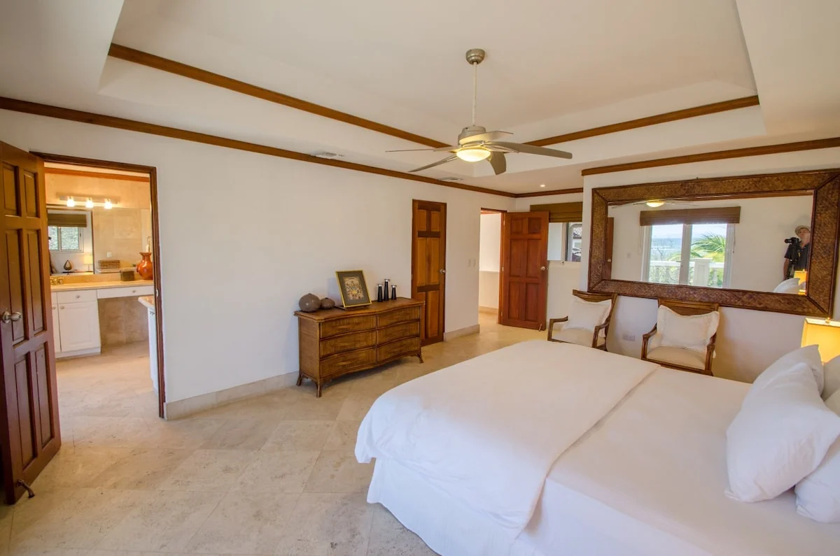 A bright bedroom with a large white bed, wooden furniture, a ceiling fan, and an open doorway toward a bathroom.