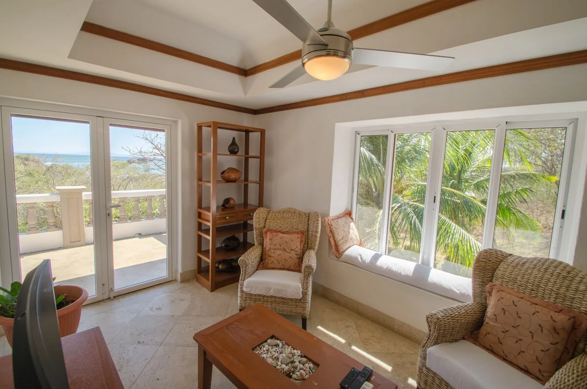 A bright living room with wicker seating, a wooden coffee table, a ceiling fan, shelving, and large windows opening toward a balcony and palm trees.