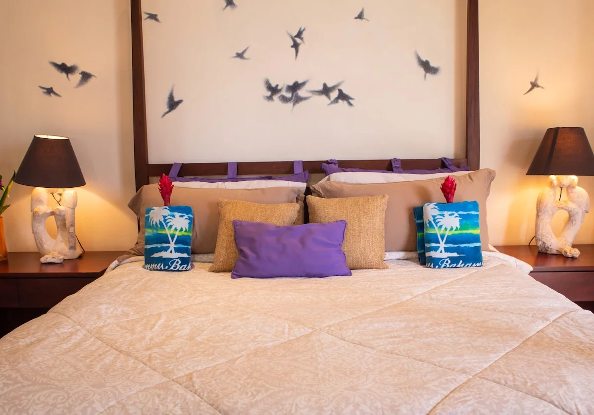 A neatly made bed with layered pillows, bedside lamps, decorative vases, and bird wall art.