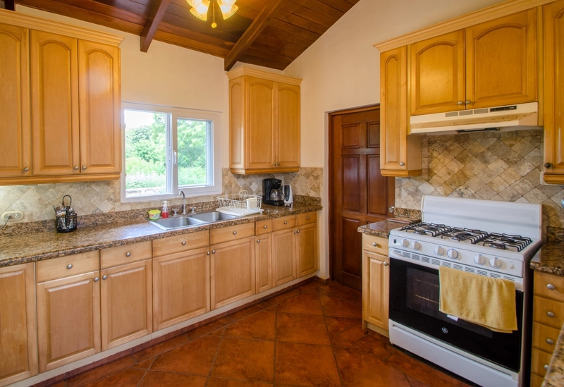 A modern kitchen featuring light wooden cabinets and granite countertops. The kitchen includes a double stainless steel sink, a white gas stove with an overhead exhaust hood, and a
