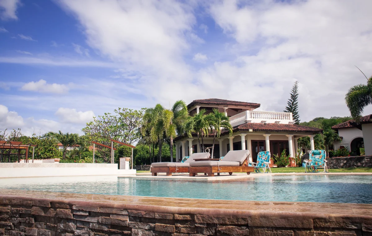 A bright swimming pool sits in front of a two-story house with palm trees, loungers, and a partly cloudy sky.