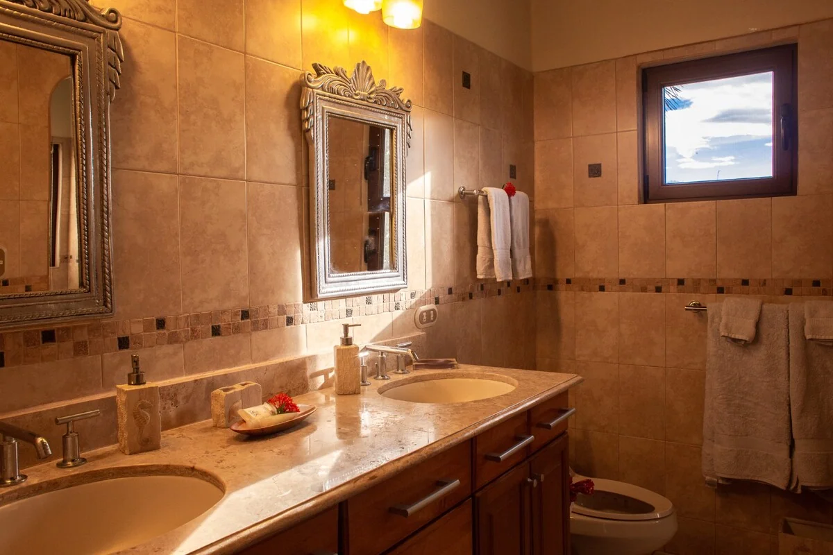 A warmly lit bathroom with two sinks, ornate mirrors, a tiled countertop, towels, a toilet, and a small window.