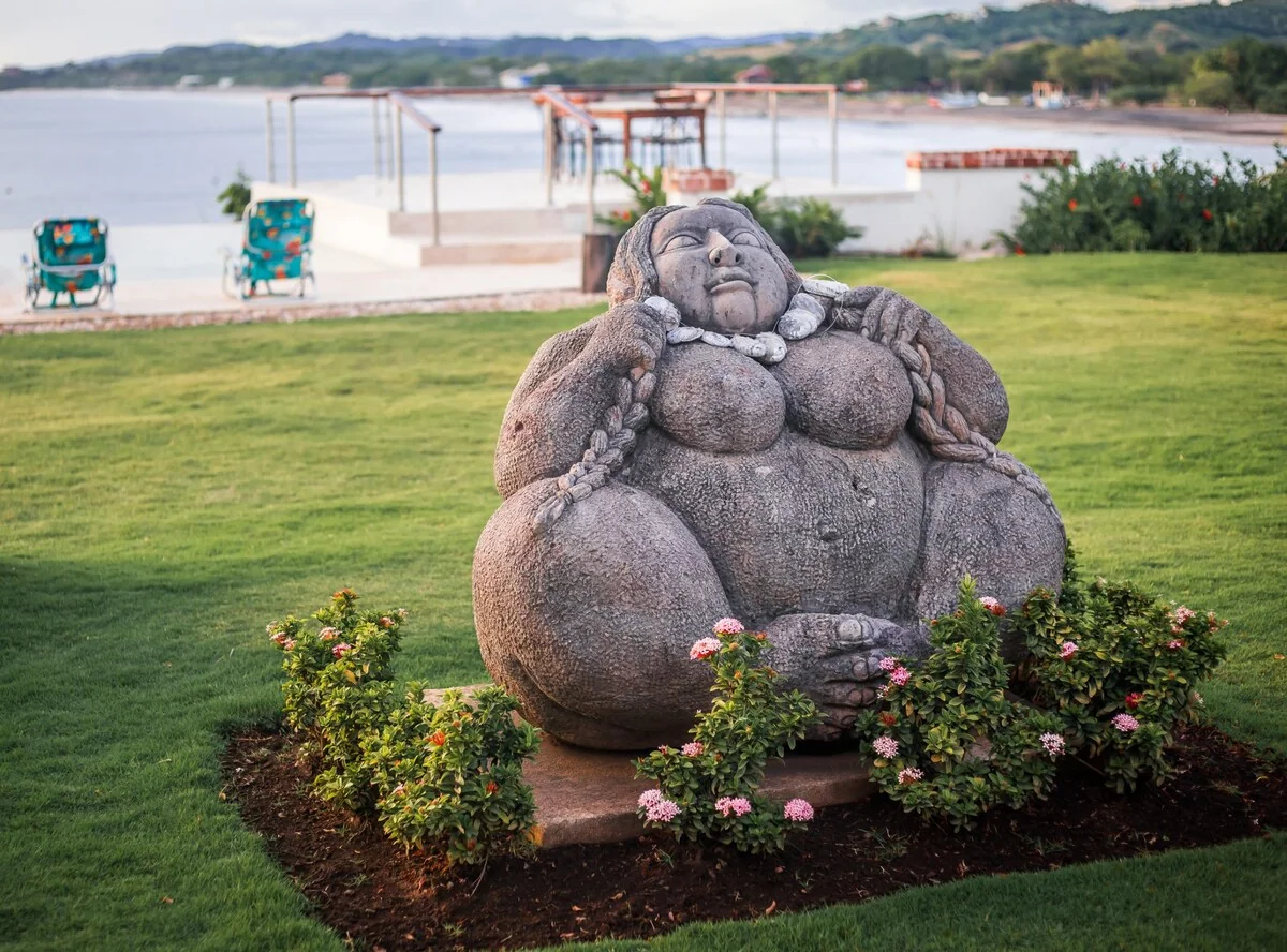 A large seated stone statue is surrounded by flowering plants on a manicured lawn beside the water.