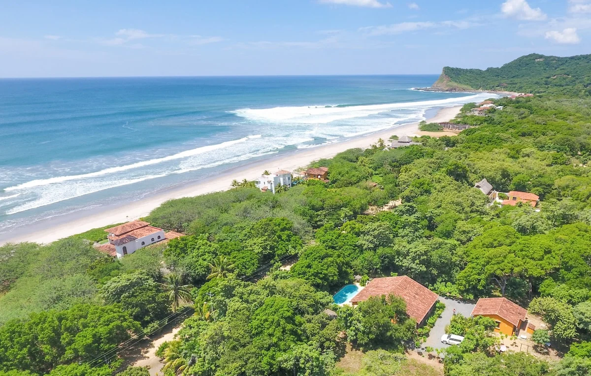 A bright aerial view shows a tropical coastline with turquoise ocean, sandy beach, dense greenery, scattered houses, and a small pool.