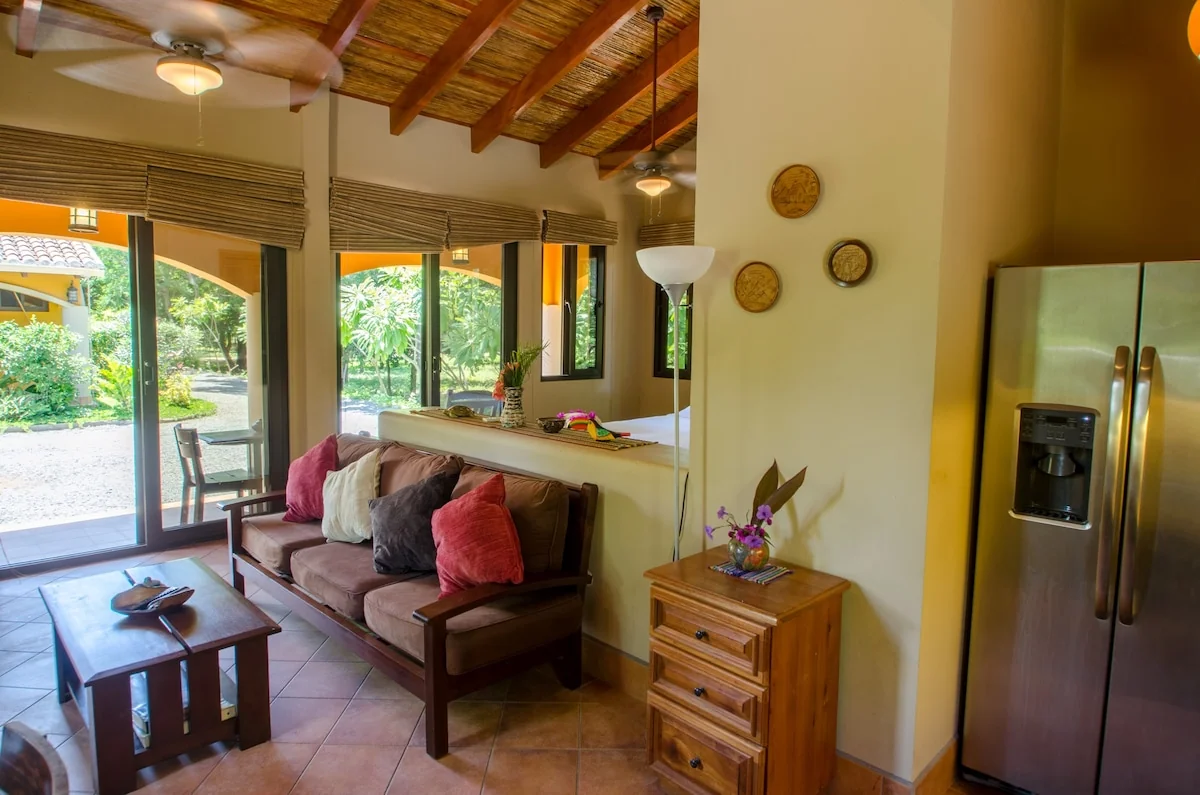 A bright living room with a sofa, coffee table, exposed wood ceiling, large windows, and a stainless steel refrigerator.