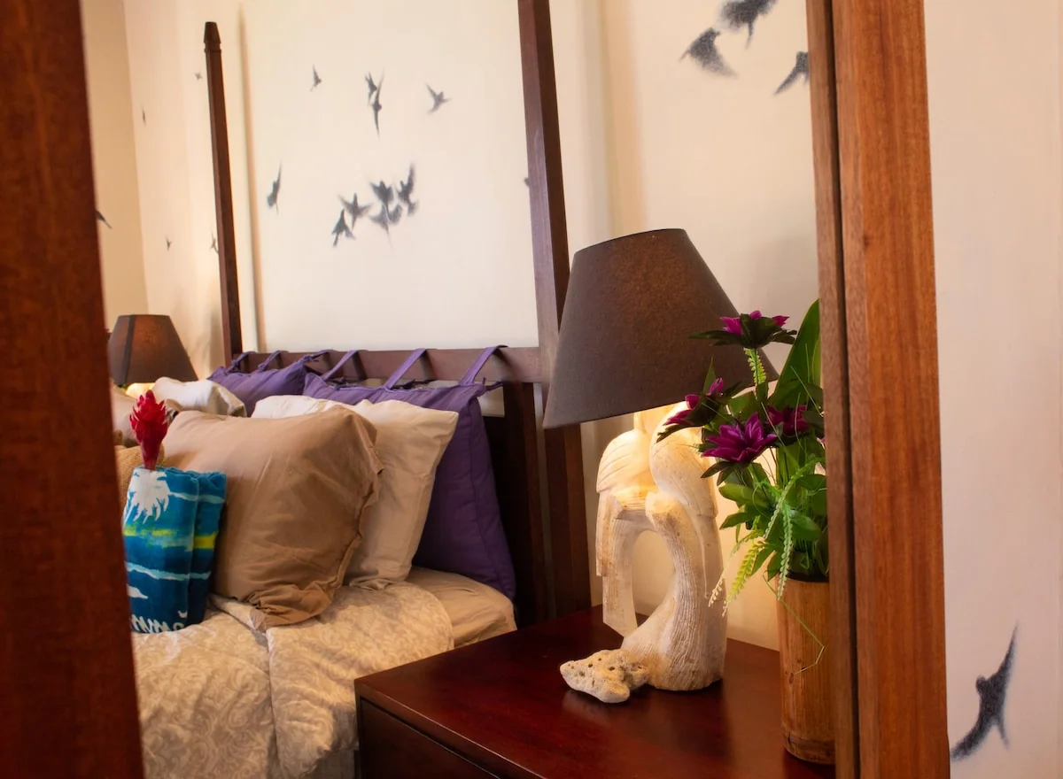 A warmly lit bedroom is reflected in a large wooden-framed mirror, showing a made bed, bedside lamp, flowers, and decorative items.