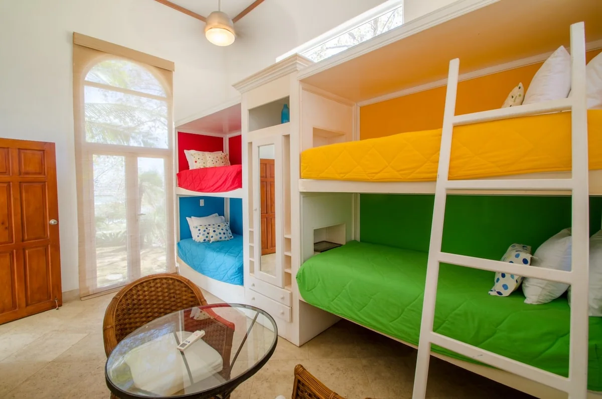 A brightly lit bedroom contains colorful bunk beds, a glass table with chairs, a wooden door, and tall windows.