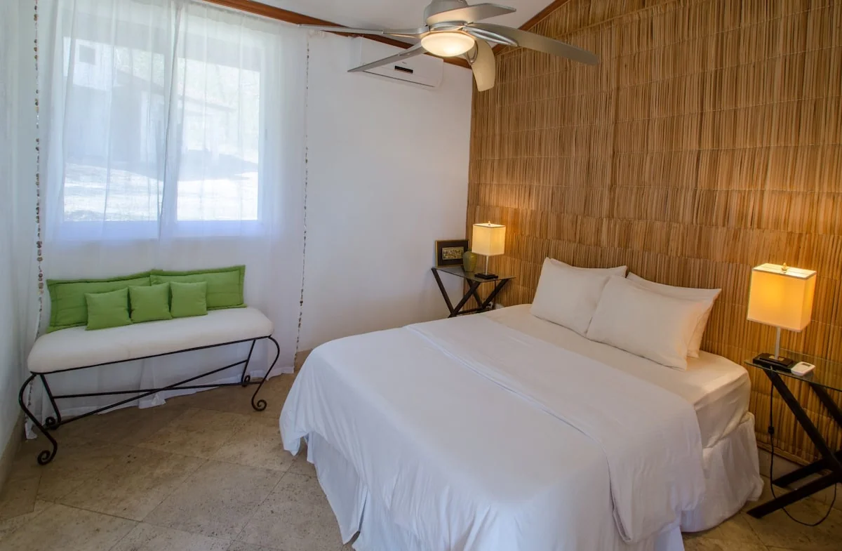 A bright bedroom with a white bed, green-cushioned bench, bedside lamps, a ceiling fan, and a textured wood wall.