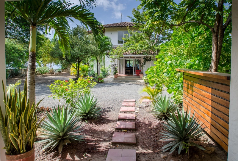 The image shows a well-maintained tropical garden with a pathway made of square stone tiles leading to the entrance of a two-story house. Various green plants and palm trees surrou