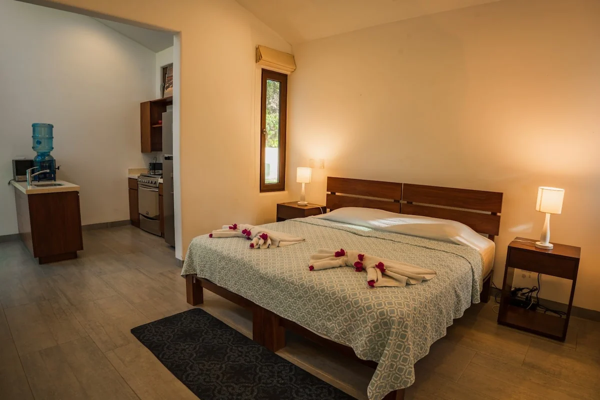 A warmly lit bedroom features a large bed with flower petals and folded towels, bedside lamps, a rug, and a water dispenser near the doorway.