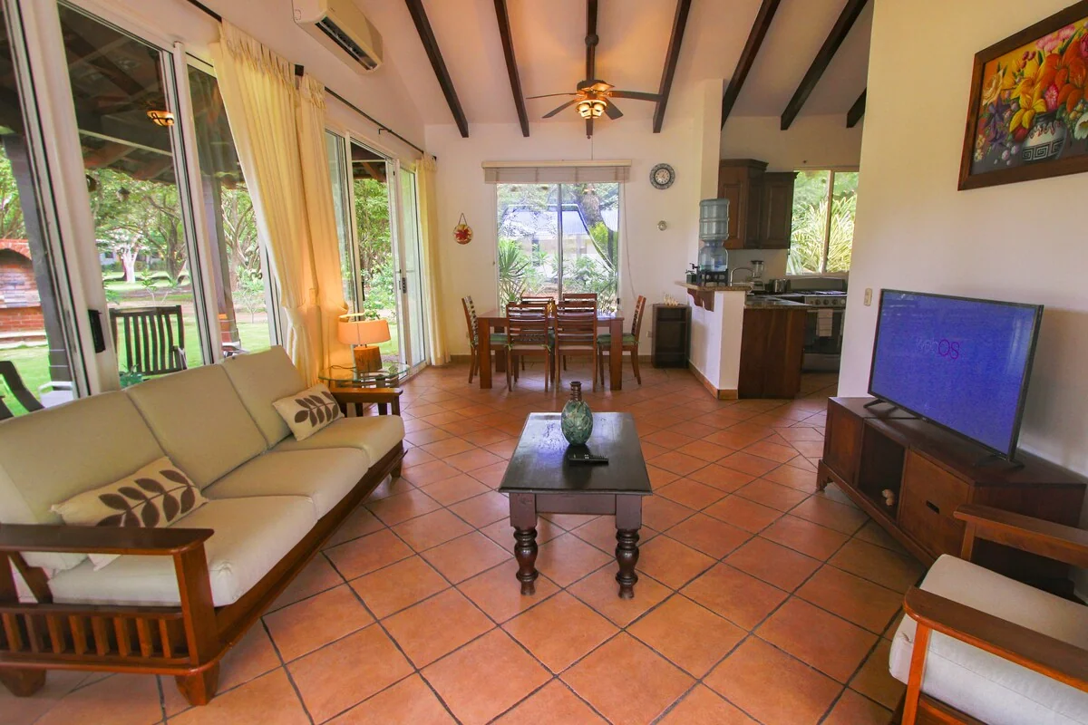 A bright open-plan living room with a sofa, coffee table, television, dining area, and visible kitchen.
