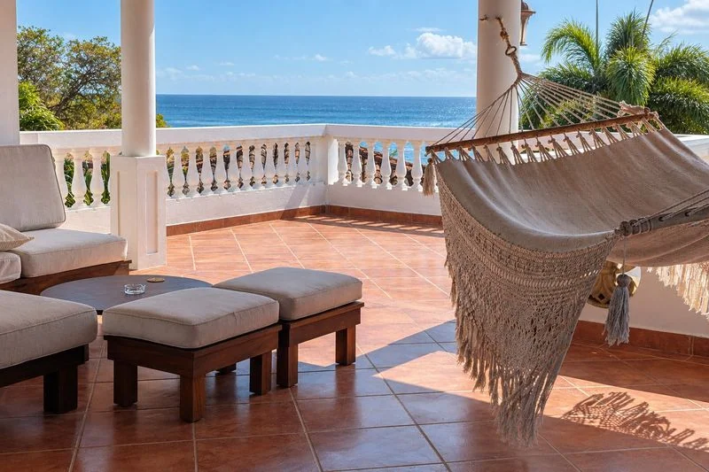 A bright tiled terrace features a fringed hammock, seating, white columns, a decorative railing, and a blue ocean beyond.