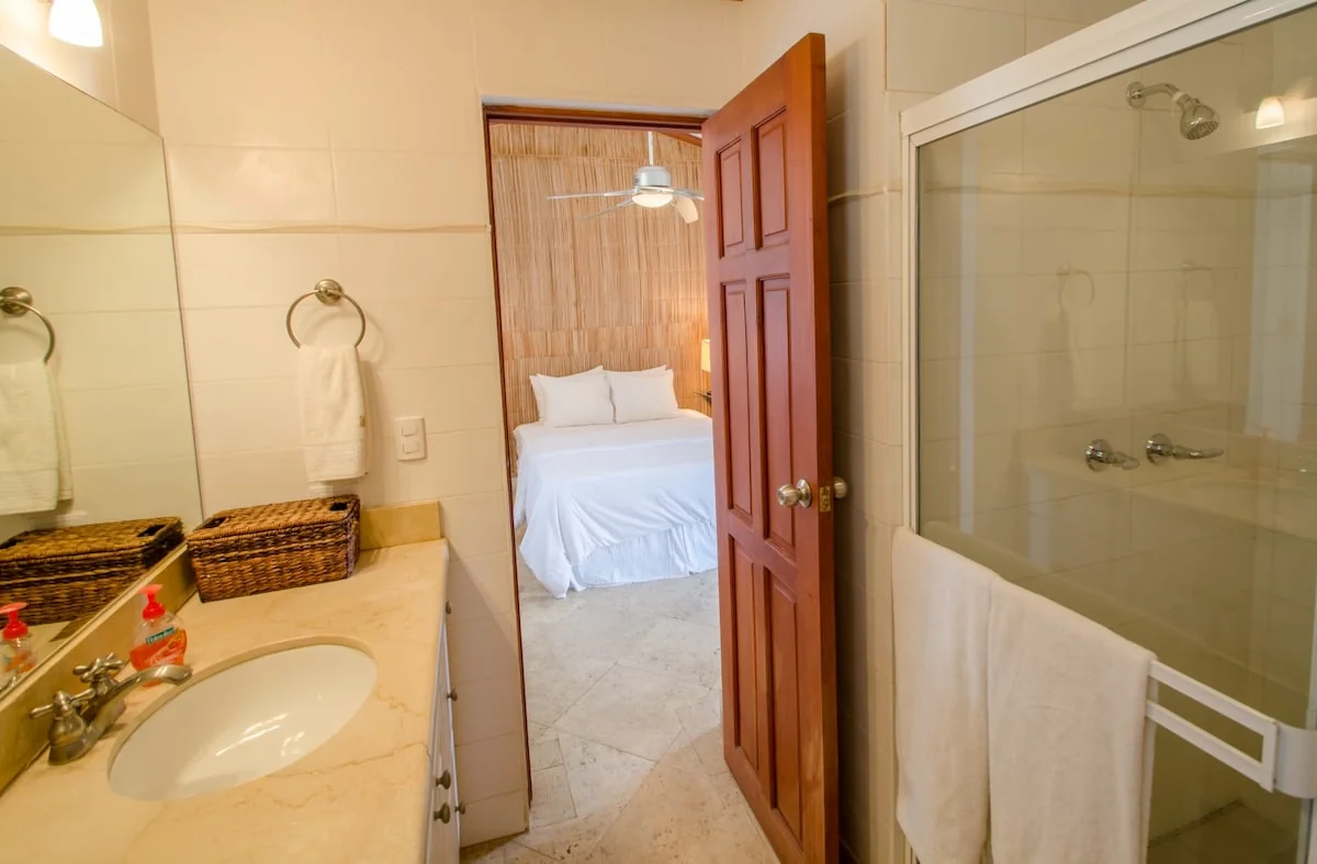 A bright bathroom with a sink, wall mirror, glass shower enclosure, towels, and an open door showing a bed beyond.