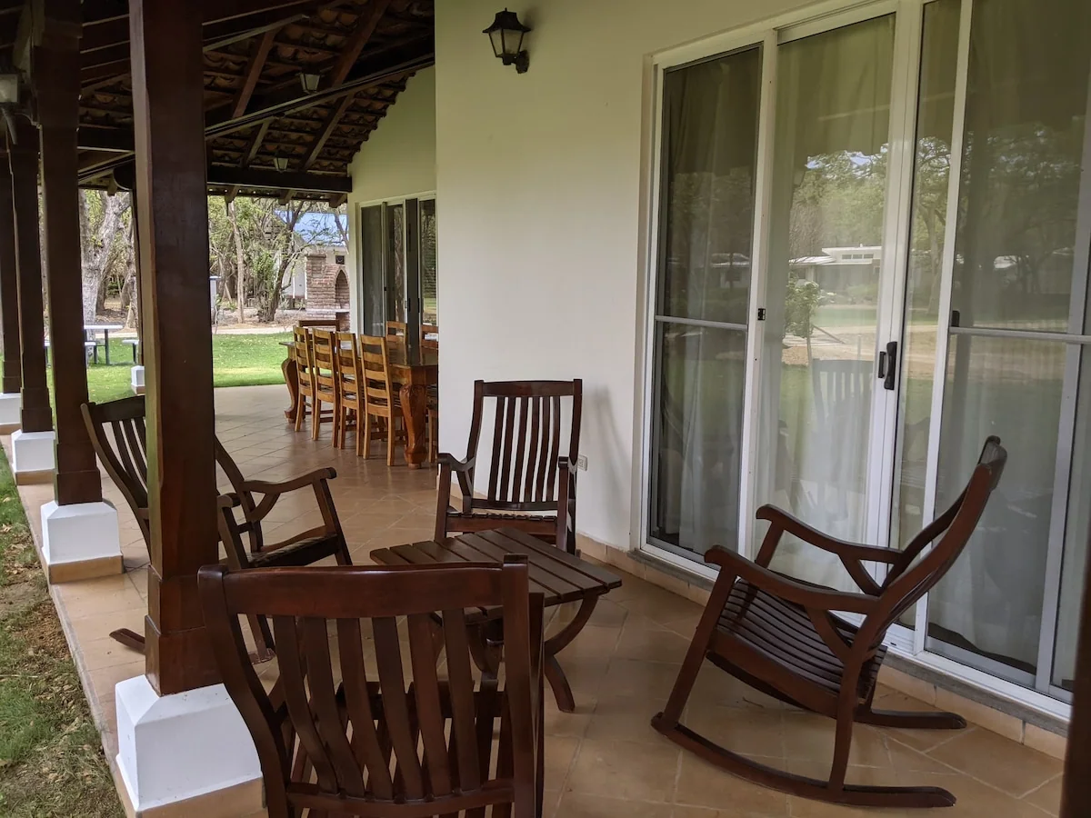 A covered terrace features several wooden rocking chairs, a small table, sliding glass doors, and a dining set extending toward the garden.