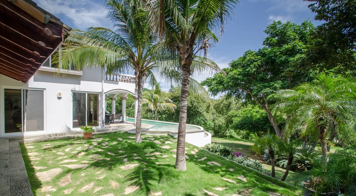 A bright landscaped garden with palm trees, a white house, and a small pool visible beside the lawn.