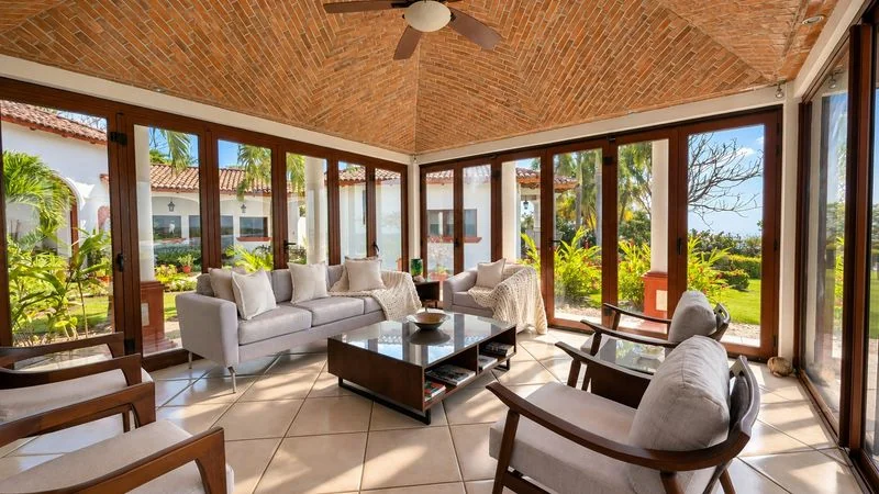 A bright glass-walled living room contains upholstered sofas and wooden armchairs around a central coffee table beneath a brick ceiling with a fan.