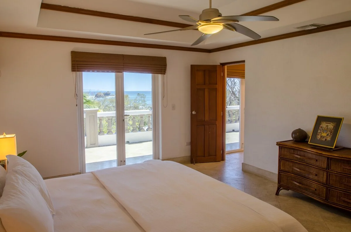 A bright bedroom with a large bed, wooden dresser, ceiling fan, and glass doors opening toward a balcony with a water view.