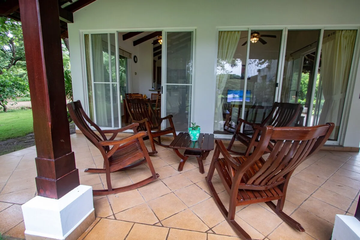 A covered terrace furnished with wooden rocking chairs and a small table overlooks a green lawn.