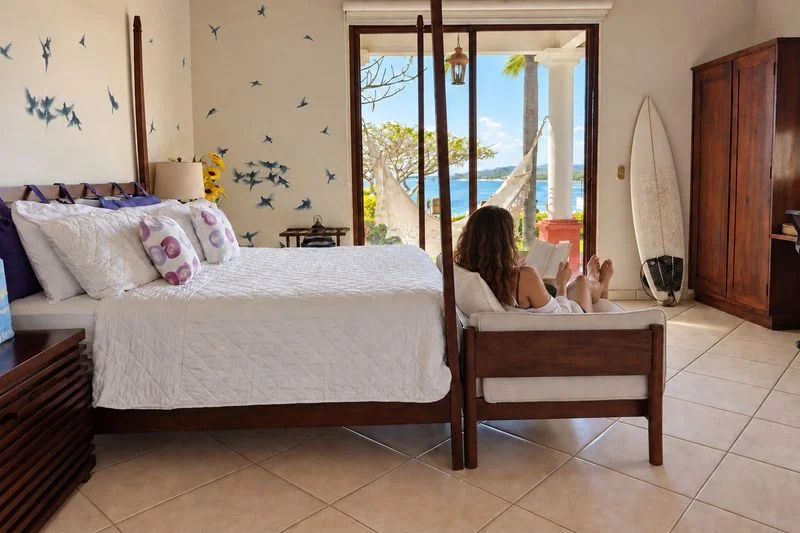 A bright bedroom with a neatly made bed, wooden furniture, bird wall decals, and a seated person beside an open window overlooking blue water.