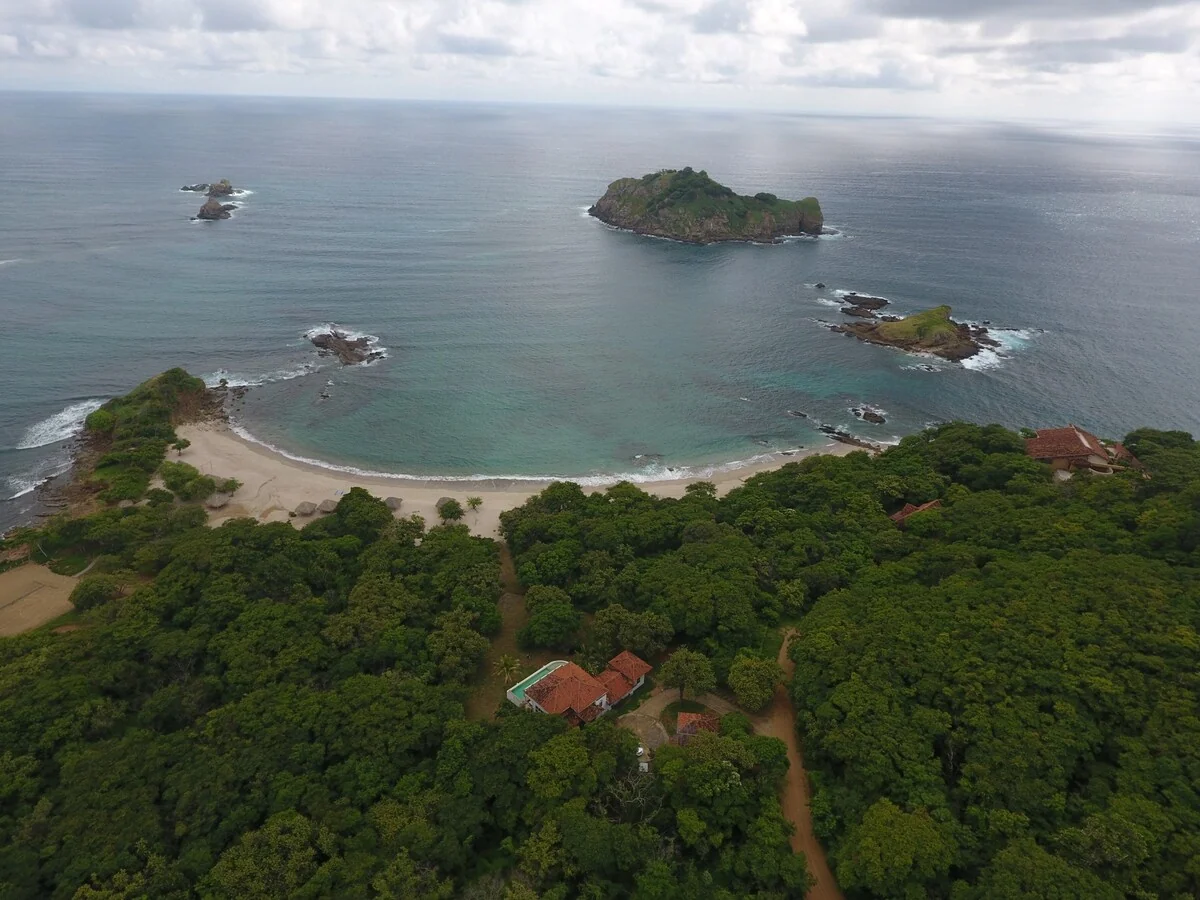 An aerial view shows a forested coastline with a curved sandy beach, rocky offshore islands, and a few red-roofed buildings beneath a cloudy sky.