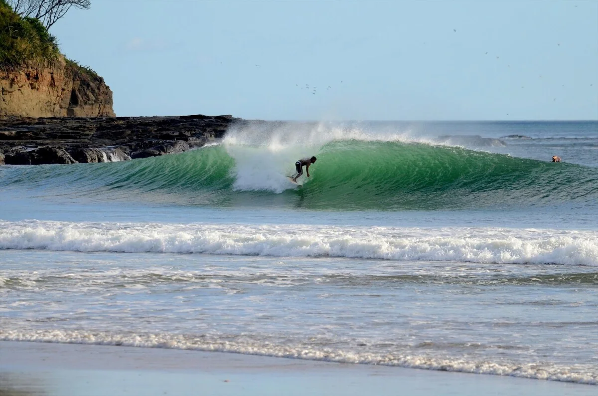 A surfer rides a breaking green wave near a rocky coastline, with another person visible farther out in the water.