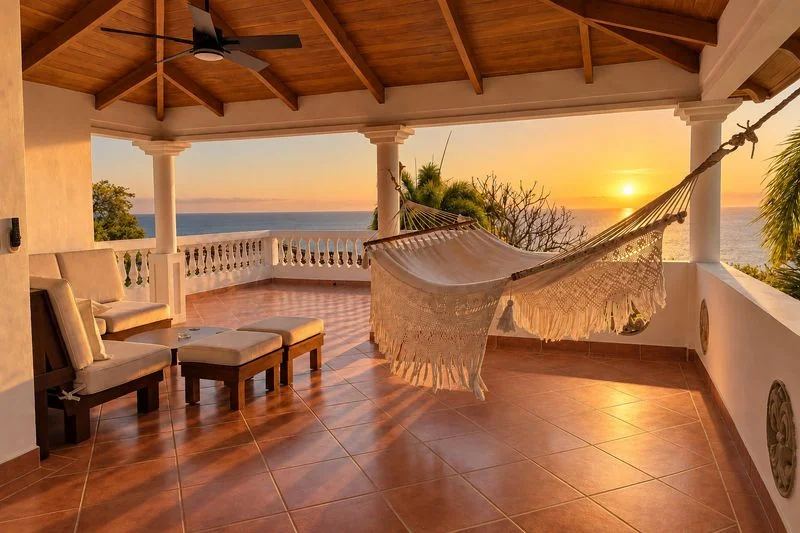 A covered tiled terrace with a hammock, seating, columns, and a sunset over the ocean.