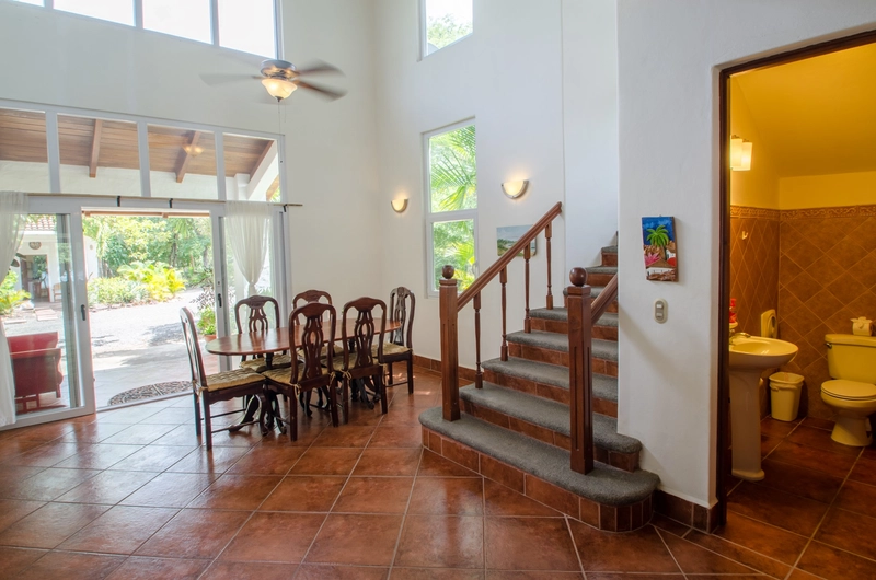 The image features a bright and open dining room with a wooden table and matching chairs. Large windows and sliding glass doors let in ample natural light, revealing views of the g