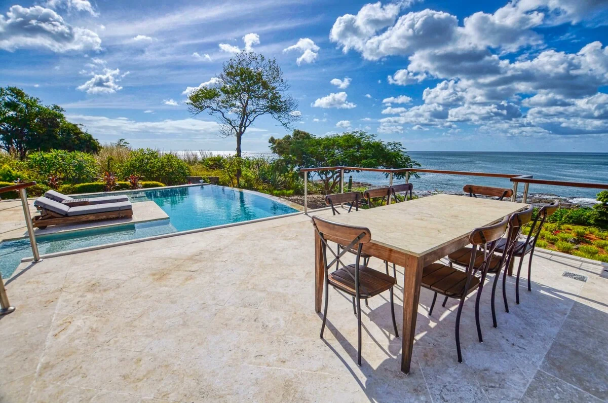 A sunny terrace overlooks a swimming pool and ocean, with a dining table and chairs in the foreground.