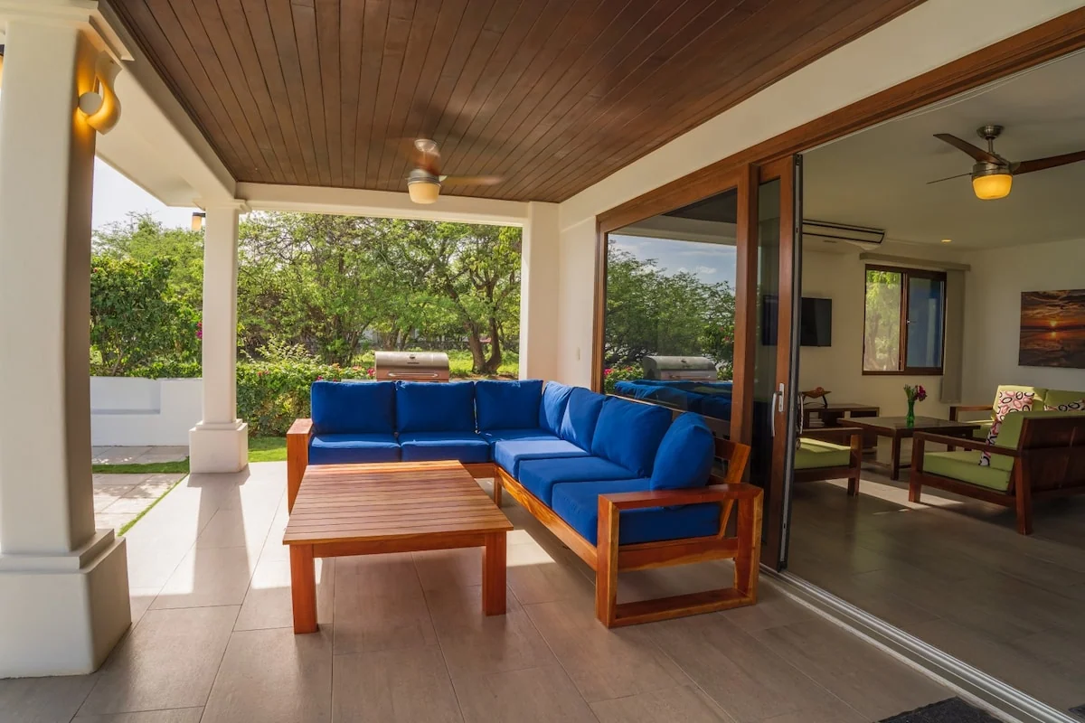 A covered terrace has a blue sectional sofa, wooden coffee table, ceiling fans, and open sliding doors leading into an interior seating area.