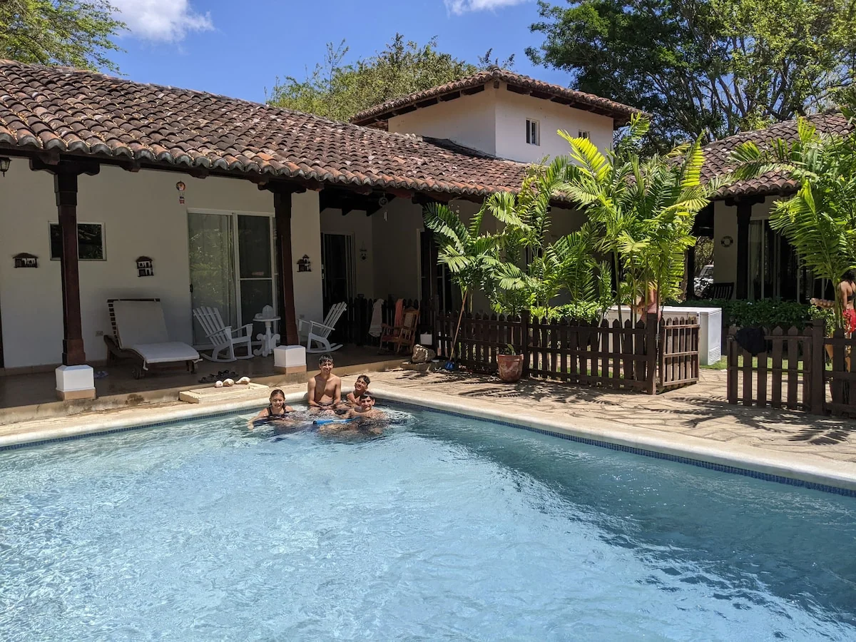 A bright swimming pool sits in front of a tiled-roof house with three people in the water and palm plants along a wooden fence.