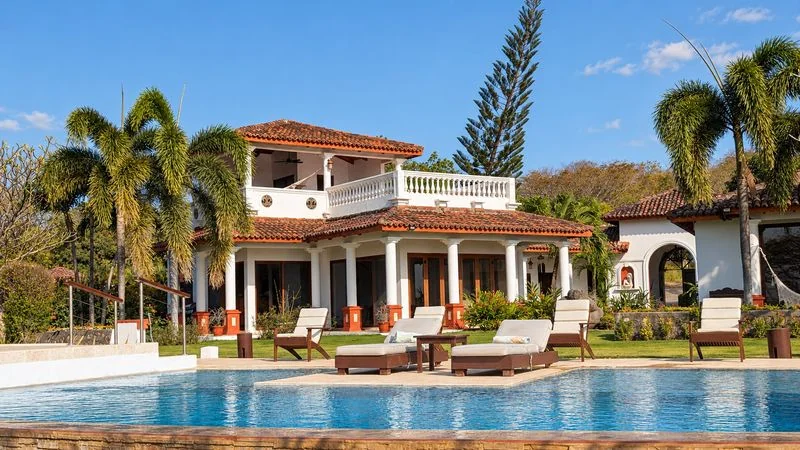 A bright blue swimming pool is set in front of a tiled-roof house with palm trees and lounge chairs.