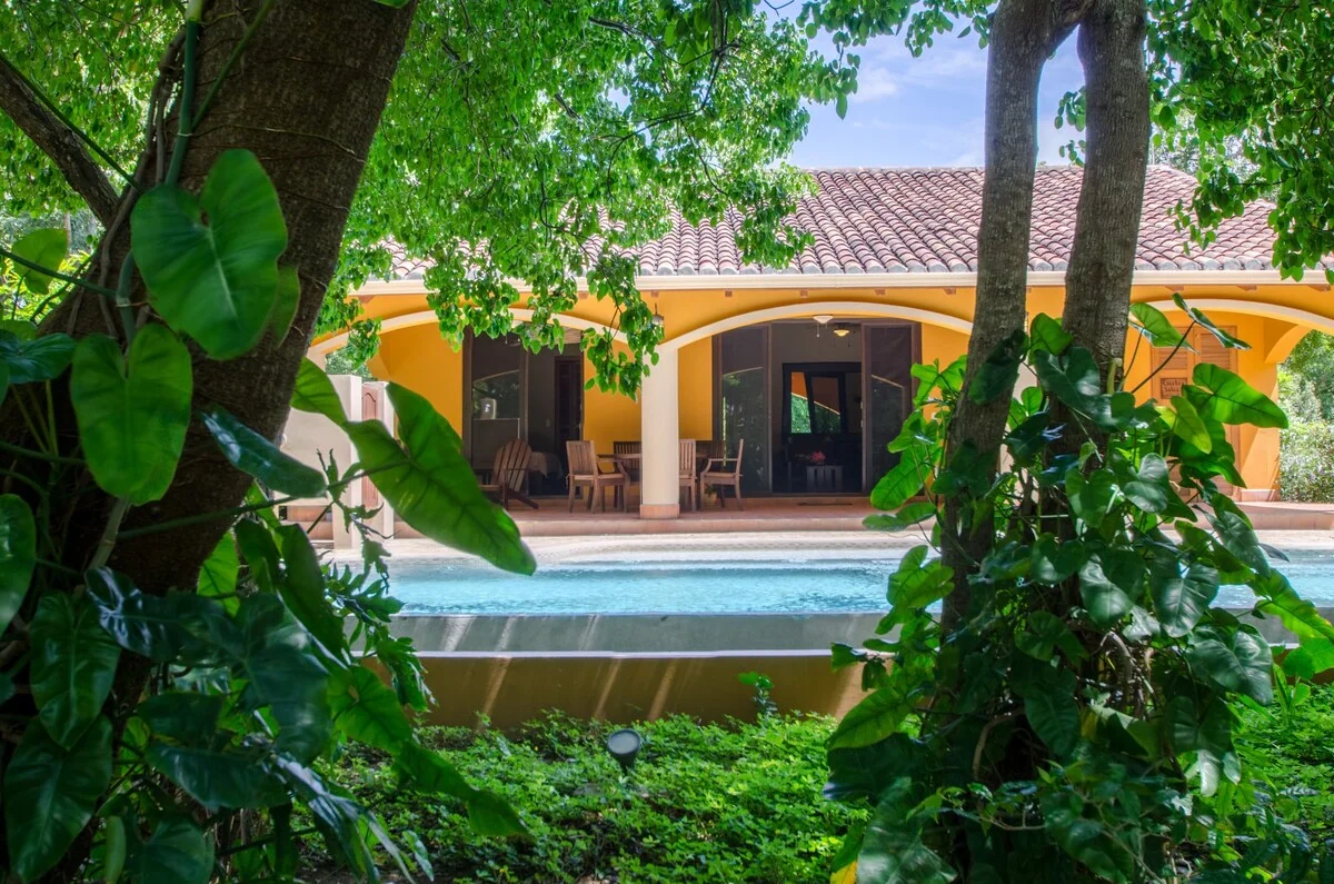 A tiled-roof yellow house with an arched patio is viewed through dense greenery across a swimming pool.