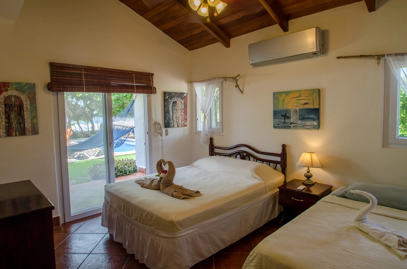 A cozy bedroom featuring a large bed with white linens and a smaller bed, both adorned with towel swans. The white walls are decorated with various paintings. A large glass door op