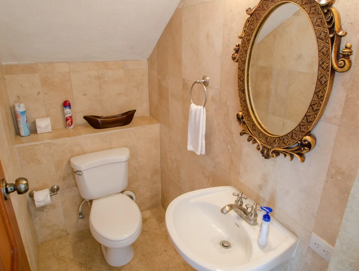 A small tiled bathroom contains a toilet, pedestal sink, ornate wall mirror, towel ring, and toiletries on a recessed shelf.