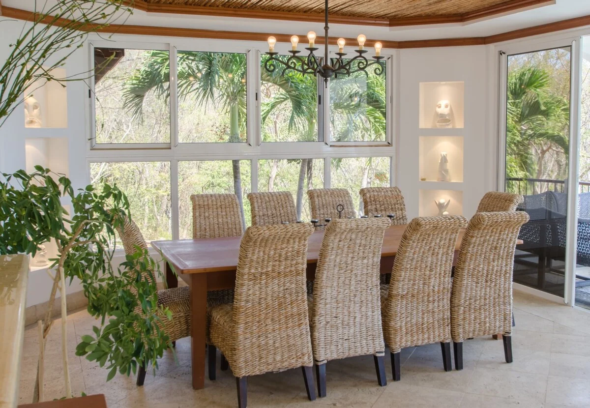 A bright dining room with a wooden table, woven chairs, chandelier, large windows, plants, and built-in display shelves.