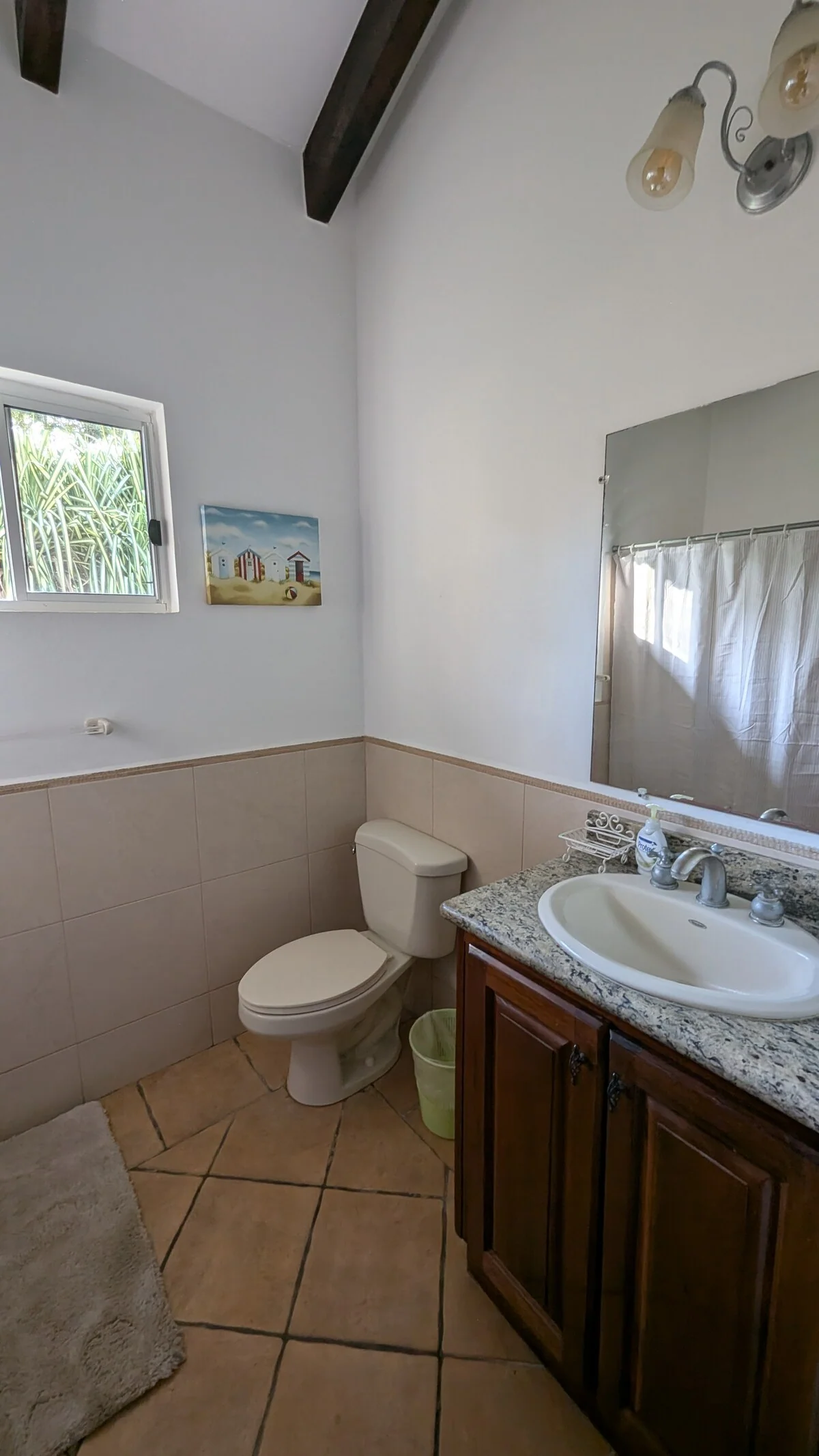 A bright bathroom contains a toilet, pedestal-style sink, wooden vanity, mirror, curtained window, and tiled walls and floor.