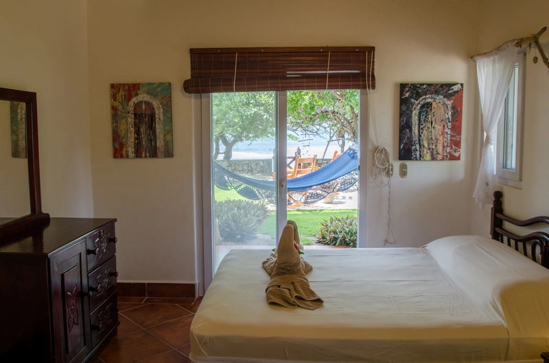 A cozy bedroom with a neatly made bed and a brown wooden dresser with a large mirror on the left side. The wall above the bed is adorned with colorful artwork. A large glass slidin