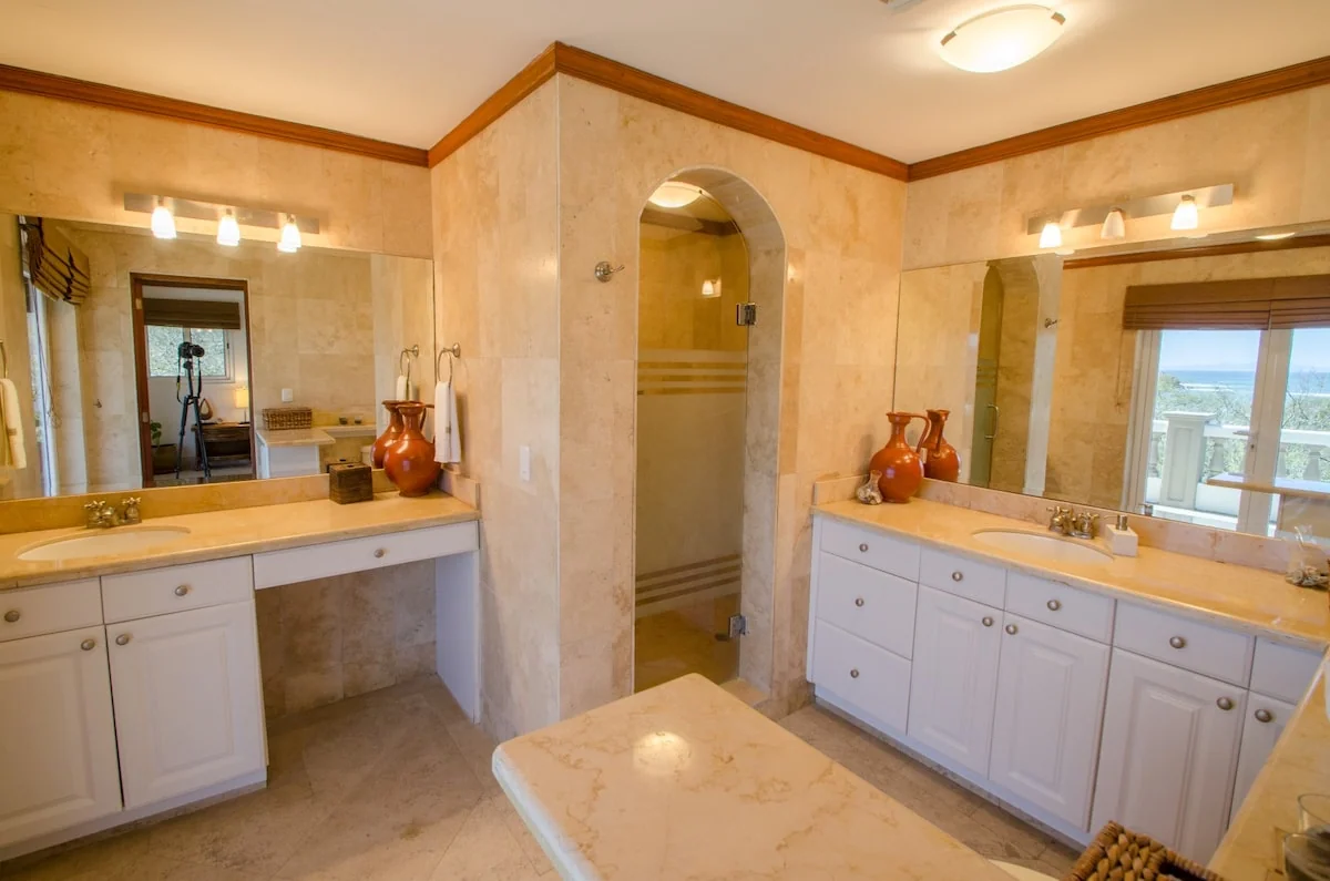 A spacious bathroom with two white vanities, large mirrors, beige stone surfaces, an arched bathtub alcove, decorative vases, and a window.