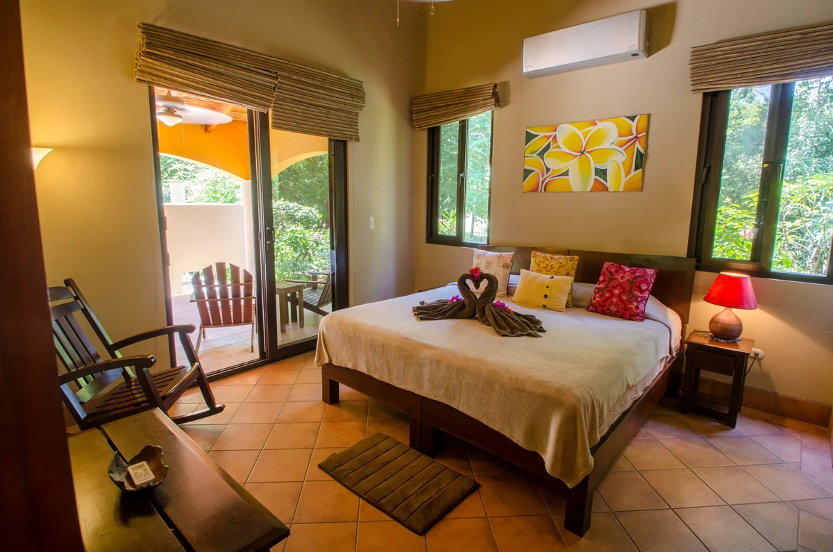A warmly furnished bedroom with a large bed, colorful pillows, wooden chairs, windows, wall artwork, and a bedside lamp.