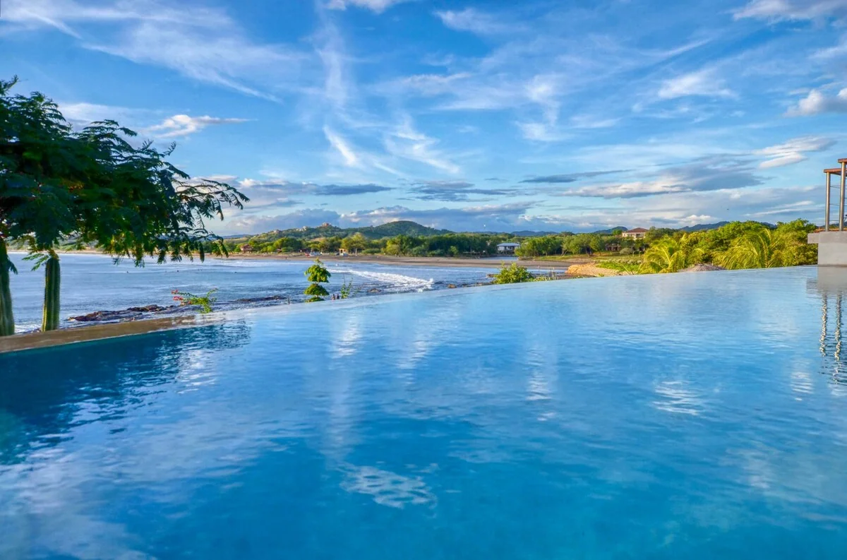 A bright blue infinity pool overlooks a shoreline, green vegetation, distant hills, and a partly cloudy sky.