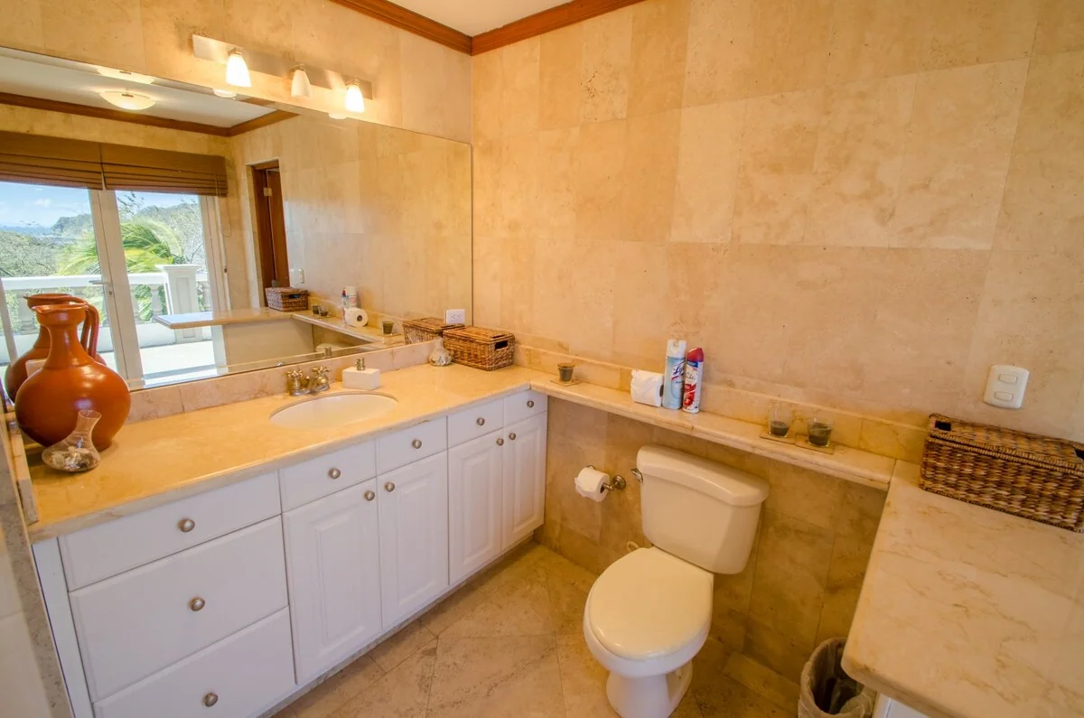 A bright bathroom with marble surfaces, a large mirror, white cabinetry, a sink, and a toilet.