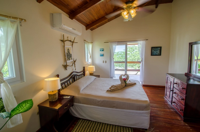 A well-lit bedroom with a wood ceiling and natural light streaming through the windows and balcony door. The room features a neatly made bed with a towel decor in the shape of swan